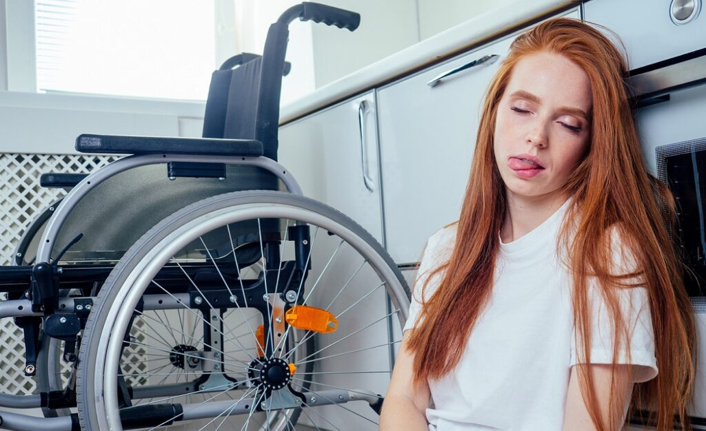Eine rothaarige junge Frau sitzt erschöpft auf dem Küchenfußboden, nachdem sie gestürzt ist. Sie leidet an Gleichgewichtsproblemen aufgrund ihrer Multiplen Sklerose (MS). Im Hintergrund steht ein Rollstuhl, der ihr als Hilfsmittel dient, aber nicht alle Herausforderungen bewältigen kann.