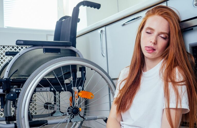 Eine rothaarige junge Frau sitzt erschöpft auf dem Küchenfußboden, nachdem sie gestürzt ist. Sie leidet an Gleichgewichtsproblemen aufgrund ihrer Multiplen Sklerose (MS). Im Hintergrund steht ein Rollstuhl, der ihr als Hilfsmittel dient, aber nicht alle Herausforderungen bewältigen kann.