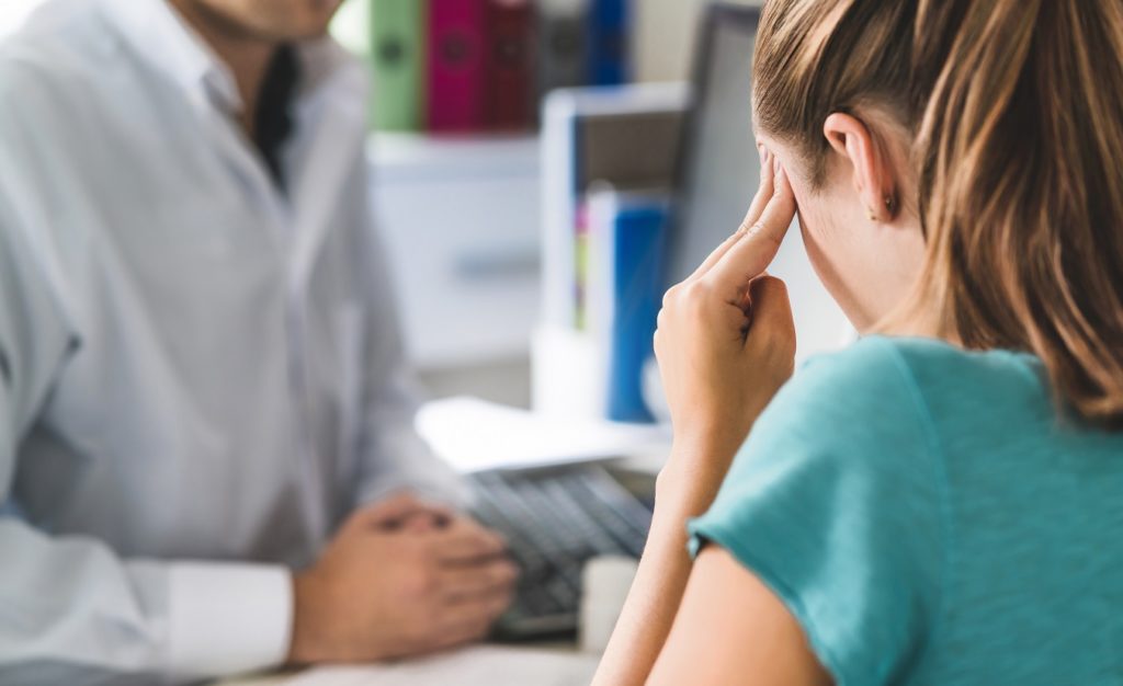Frau leidet unter starken Kopfschmerzen als Symptom der Multiplen Sklerose und sitzt bei einem Arzt im Sprechzimmer.