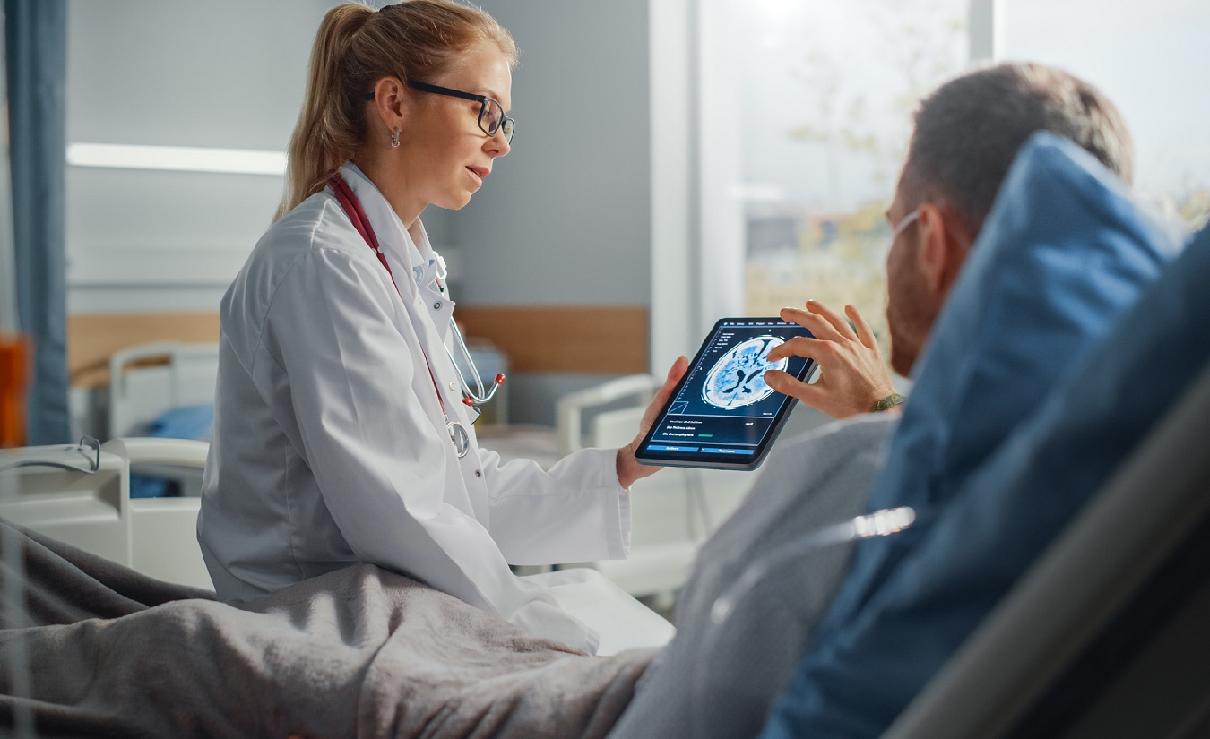 Ein kranker Mann liegt im Bett und spricht mit einer freundlichen Ärztin, die ein Tablet-Computer benutzt, um mit ihm zu kommunizieren.