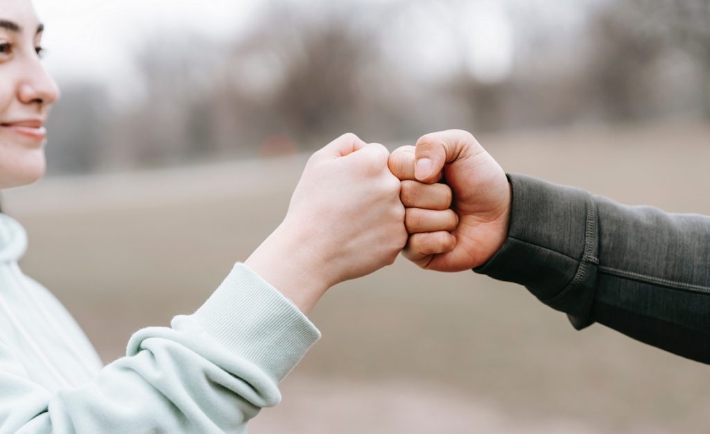 Mann und Frau würdigen sich anerkennend mit einem Handshake. Die Frau lächelt dabei und trägt einen hellen Pullover, während der Mann einen dunklen Pullover trägt.