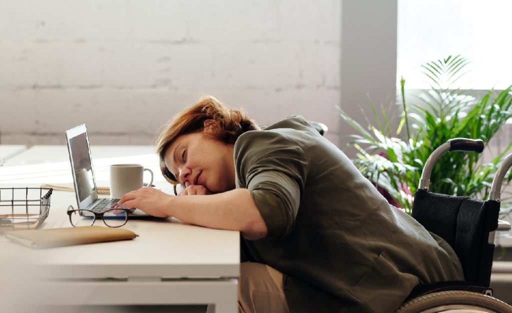 Eine Frau mit MS leidet unter Erschöpfung und hat ihren Kopf seitlich auf den Schreibtisch gelegt. Sie sitzt im Rollstuhl und arbeitet an ihrem Laptop. Neben dem Laptop stehen eine Kaffeetasse und liegt eine Brille. Eine grüne Zimmerpflanze ist im Hintergrund zu sehen.