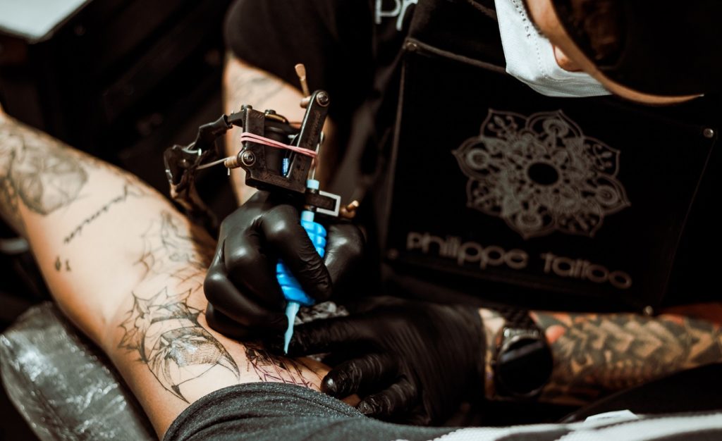 Ein Tätowierer mit Maske und Tattoos an den Armen tätowiert den Unterarm eines Kunden.