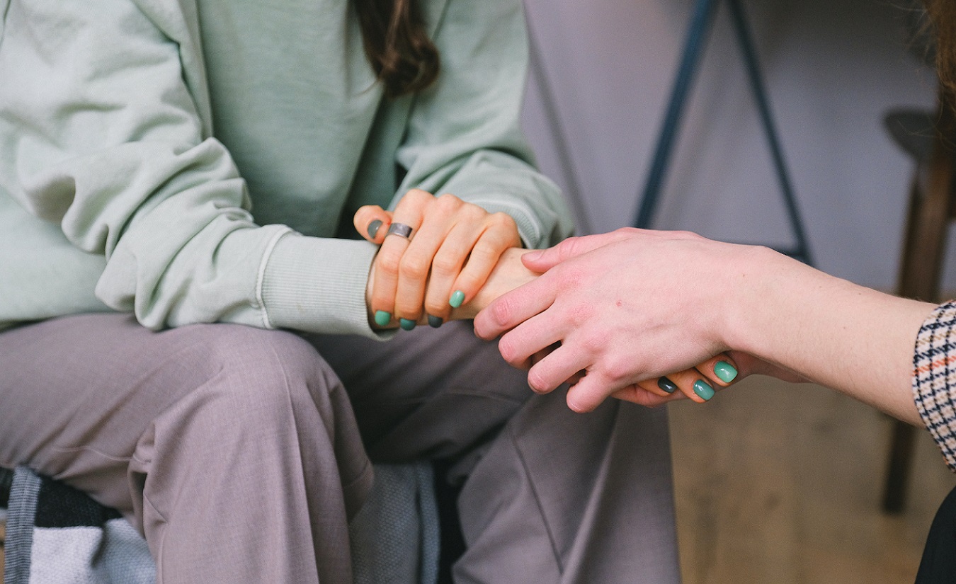 Eine jüngere Frau mit dunklem, langem Haar und grünen Fingernägeln wird von einer anderen Person getröstet und gehalten.