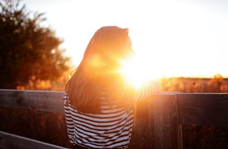 Eine Frau mit Multipler Sklerose (MS) steht auf einem Balkon und betrachtet den eindrucksvollen Sonnenuntergang über einem Bauernhof.