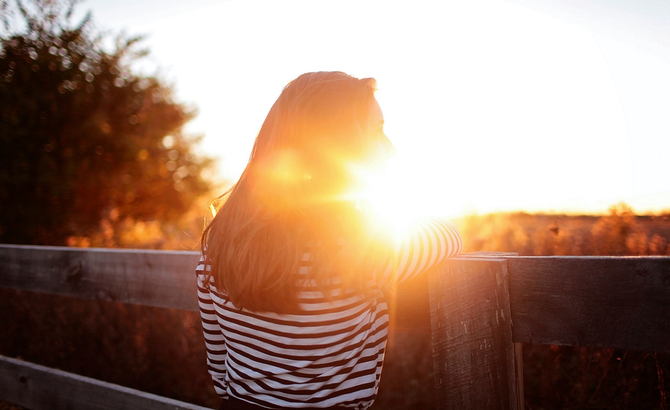 Eine Frau mit Multipler Sklerose (MS) steht auf einem Balkon und betrachtet den eindrucksvollen Sonnenuntergang über einem Bauernhof.