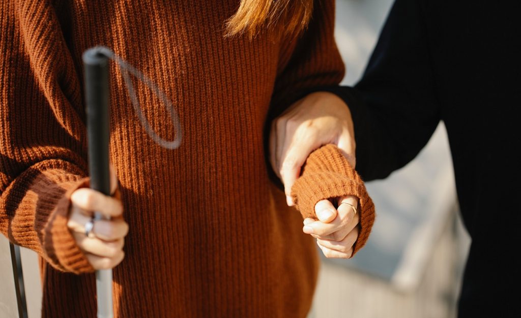 Eine Frau mit Sehbehinderung nutzt einen Stock und wird von einer anderen Person gehalten. Das Bild betont die Bedeutung von Hilfsmitteln zur Linderung von MS-Symptomen.