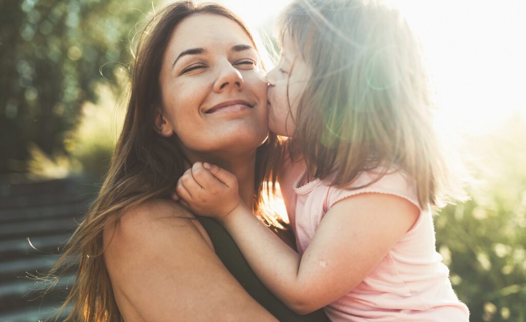 Eine Mutter, die trotz ihrer Multiplen Sklerose (MS) einen erfüllten Kinderwunsch erlebt, wird von ihrer Tochter liebevoll auf die Wange geküsst. Ein Bild des Glücks in der Natur.