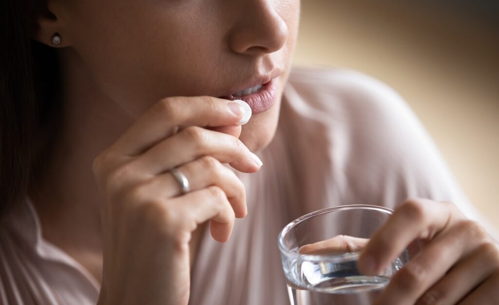 Junge Frau hält eine Tablette an ihren Mundwinkel und hält ein Glas Wasser in der Hand.