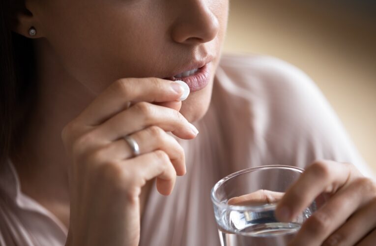 Junge Frau hält eine Tablette an ihren Mundwinkel und hält ein Glas Wasser in der Hand.