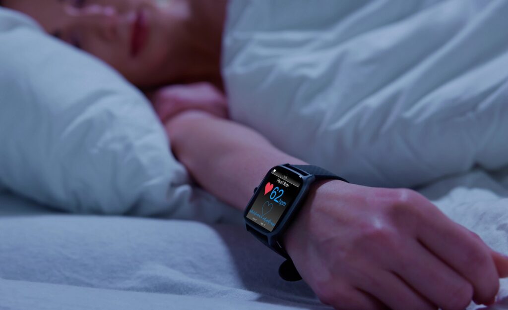 Eine Frau mit Multipler Sklerose (MS) schläft in einem hellblauen Bett. Sie trägt eine Uhr, die ihre Schlafstörungen und Herzfrequenz überwacht. Die Frau hält ihre rechte Hand aus der Bettdecke, wo man auch die Uhr sieht.