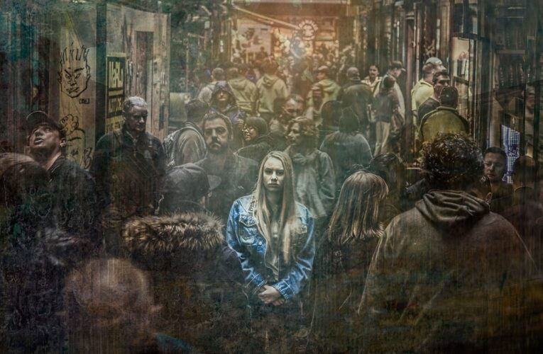 Ein farbiges Porträt einer jungen Frau inmitten einer schwarz-weißen Menschenmenge. Die Frau mit blonden Haaren und einer blauen Jeansjacke wirkt überfordert und ängstlich, möglicherweise aufgrund einer Angststörung in Verbindung mit oder zusätzlich zur Multiplen Sklerose.
