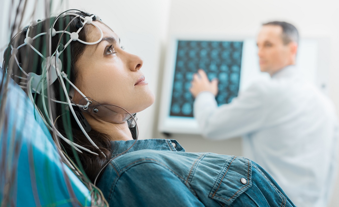 Eine entspannte Frau mit Multipler Sklerose (MS) unterzieht sich einer Elektroenzephalographie (EEG)-Untersuchung. Ihr Kopf ist verkabelt, während im Hintergrund ein aufmerksamer Arzt die Untersuchung überwacht. Die Frau mit langen schwarzen Haaren ist dezent geschminkt und trägt ein blaues Jeanshemd.