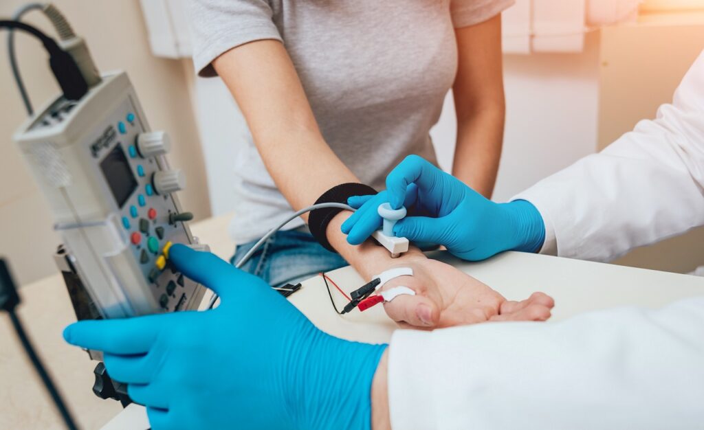 Medizinische Fachangestellte führt einen Nervenleitgeschwindigkeitstest an einer Frau mit MS durch. Elektrische Stimulation am Unterarm und Aufzeichnung der Impulse über Klebeelektroden.