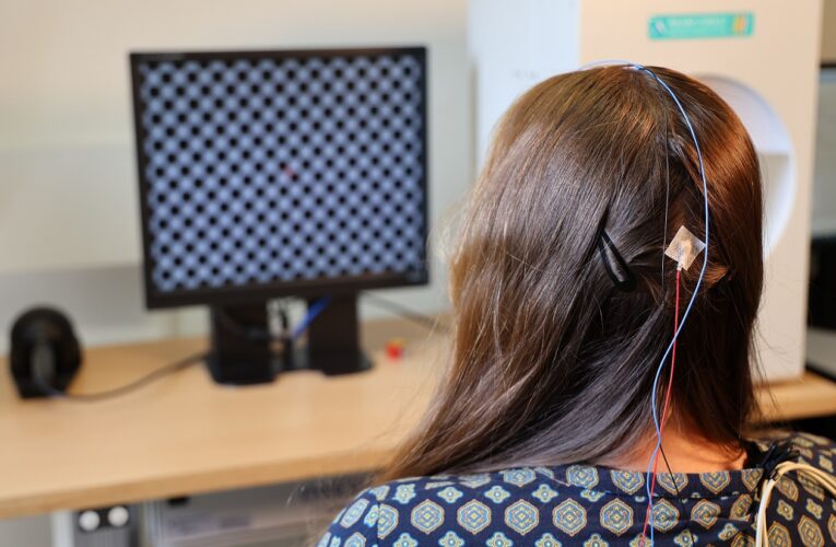 Eine junge Frau mit langen Haaren sitzt konzentriert vor einem Bildschirm, während sie an einer visuell evozierten Potenzial (VEP)-Untersuchung teilnimmt. Dabei werden Reize von einem schachbrettartigen Muster gemessen. Elektroden sind am Hinterkopf der Frau angebracht.