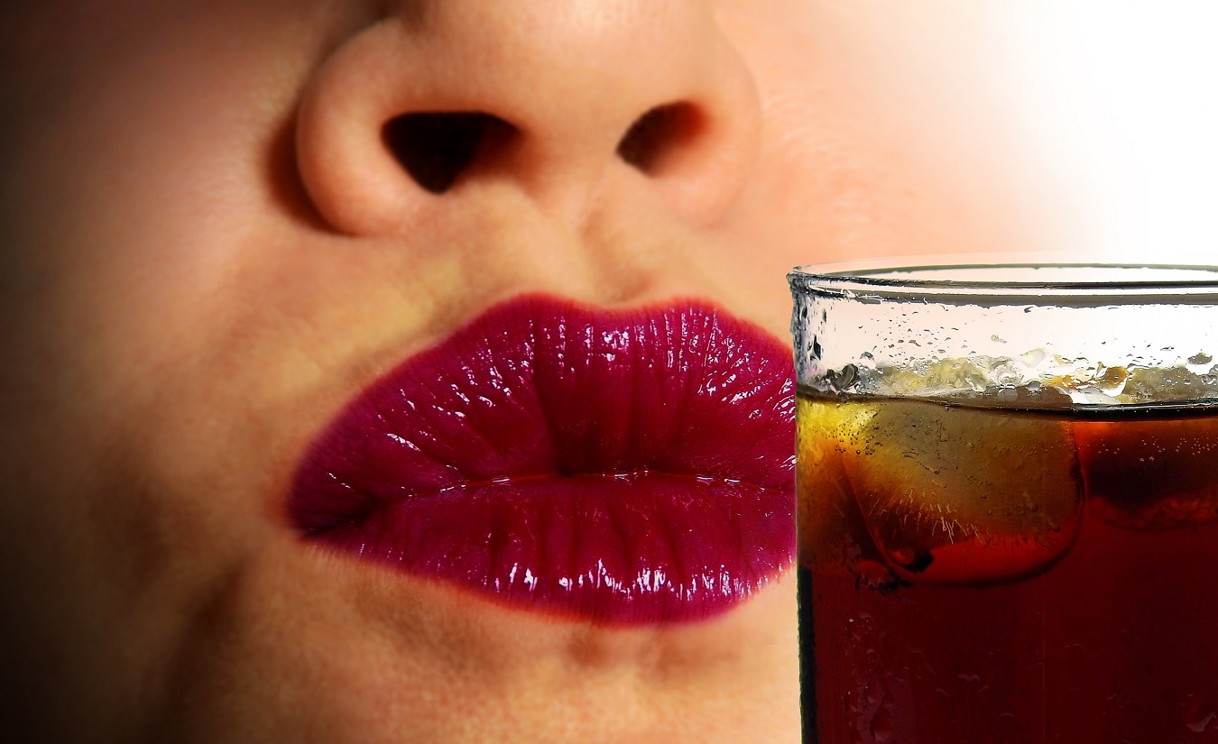 Nahaufnahme einer Frau, die ein bräunliches Getränk mit Aspartam trinkt. Ihre Lippen sind leicht angefeuchtet, sie trägt roten Lippenstift.