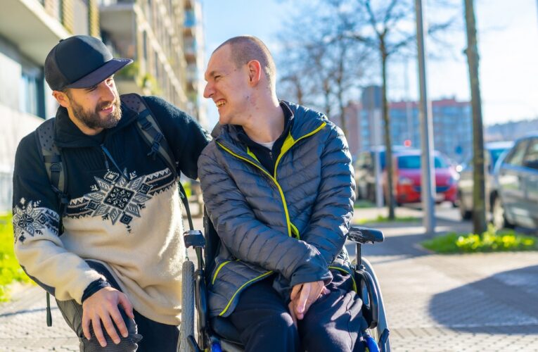 Ein Mann im Rollstuhl auf einem Gehweg wird von seinem Freund begleitet, der neben dem Rollstuhl kniet. Die beiden schauen sich lachend an, ein Symbol für Lebensfreude trotz MS.