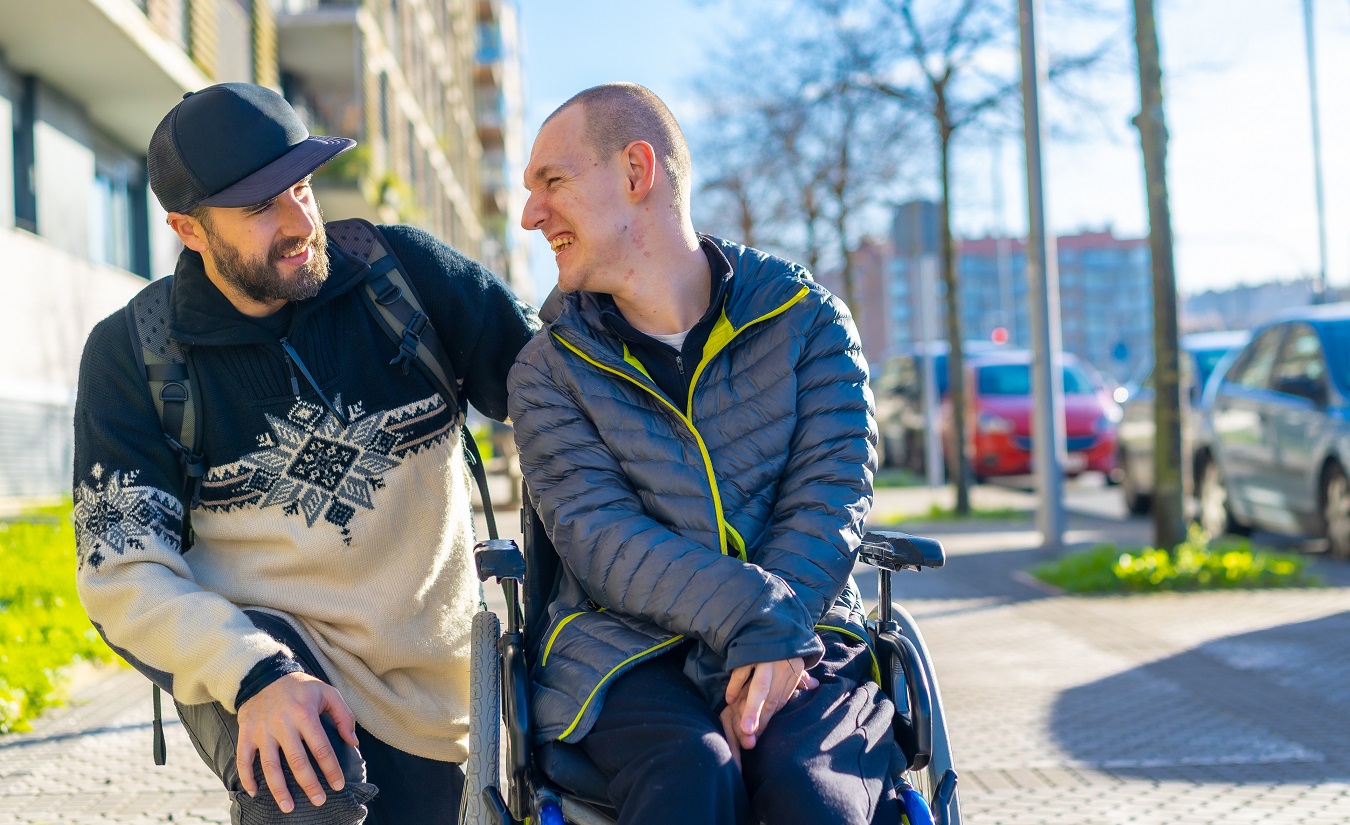 Ein Mann im Rollstuhl auf einem Gehweg wird von seinem Freund begleitet, der neben dem Rollstuhl kniet. Die beiden schauen sich lachend an, ein Symbol für Lebensfreude trotz MS.