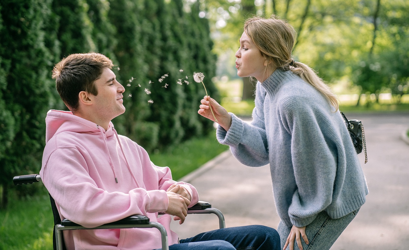 Ein glückliches Paar, bestehend aus einem Mann im Rollstuhl und einer Frau, die ihm verliebt eine Pusteblume ins Gesicht bläst, während sie gemeinsam die Natur genießen.