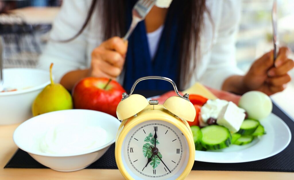 Eine Frau sitzt an einem Tisch und betrachtet eine Schüssel mit frischem Obst und Gemüse neben einem gelben Wecker.