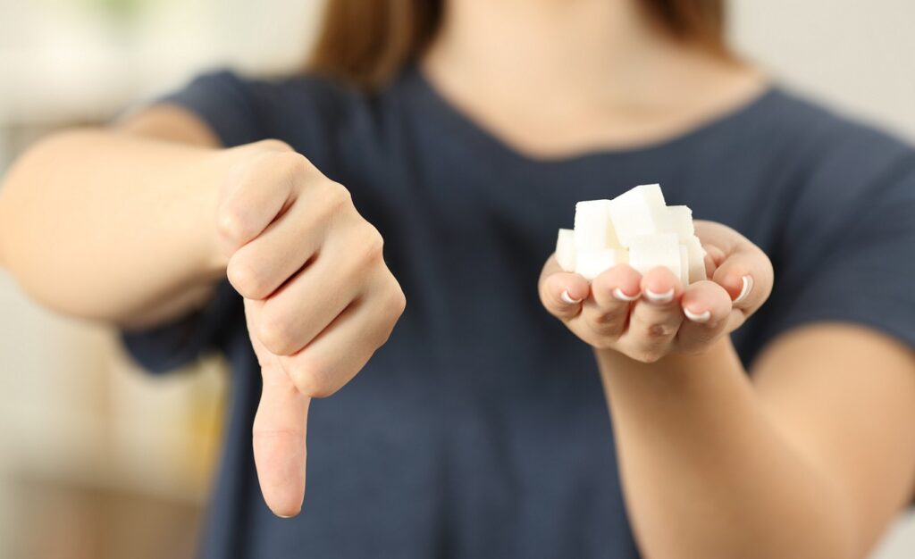Eine Frau hält einen Würfelzucker in ihrer linken Hand und zeigt mit ihrer rechten Hand den Daumen nach unten.