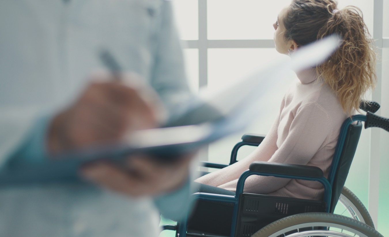 Ein Arzt schreibt Krankenakten, während ein Patient im Rollstuhl wartet.