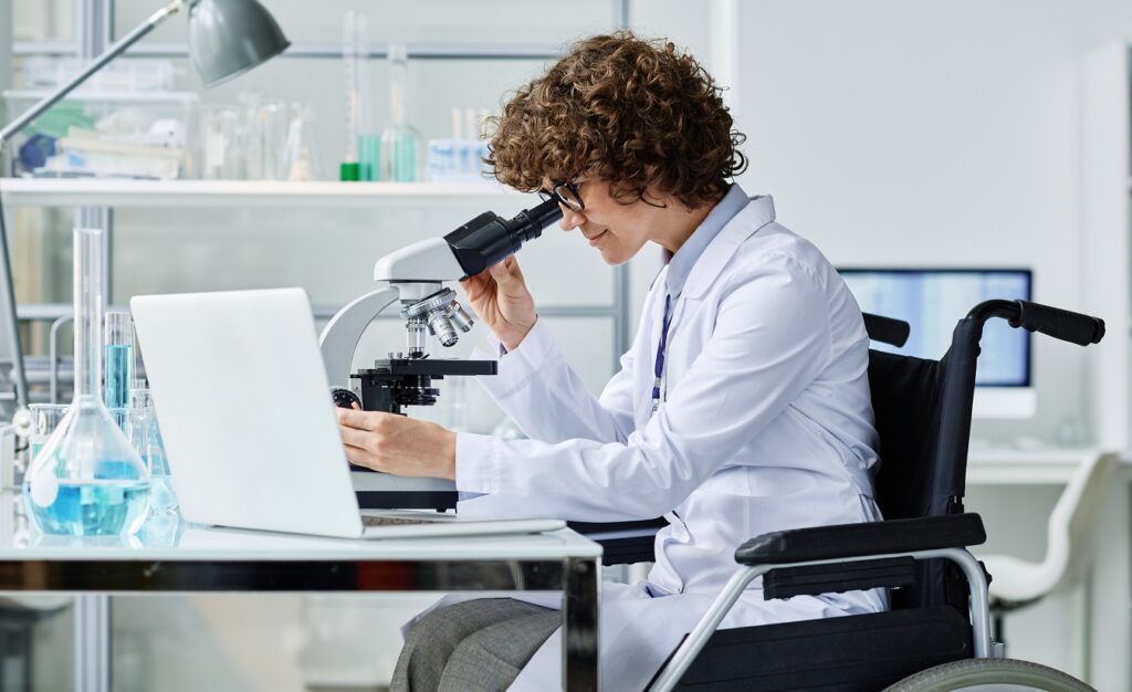 Eine junge Frau mit MS sitzt im Rollstuhl in einem Labor, während sie konzentriert an einem Arbeitstisch arbeitet.