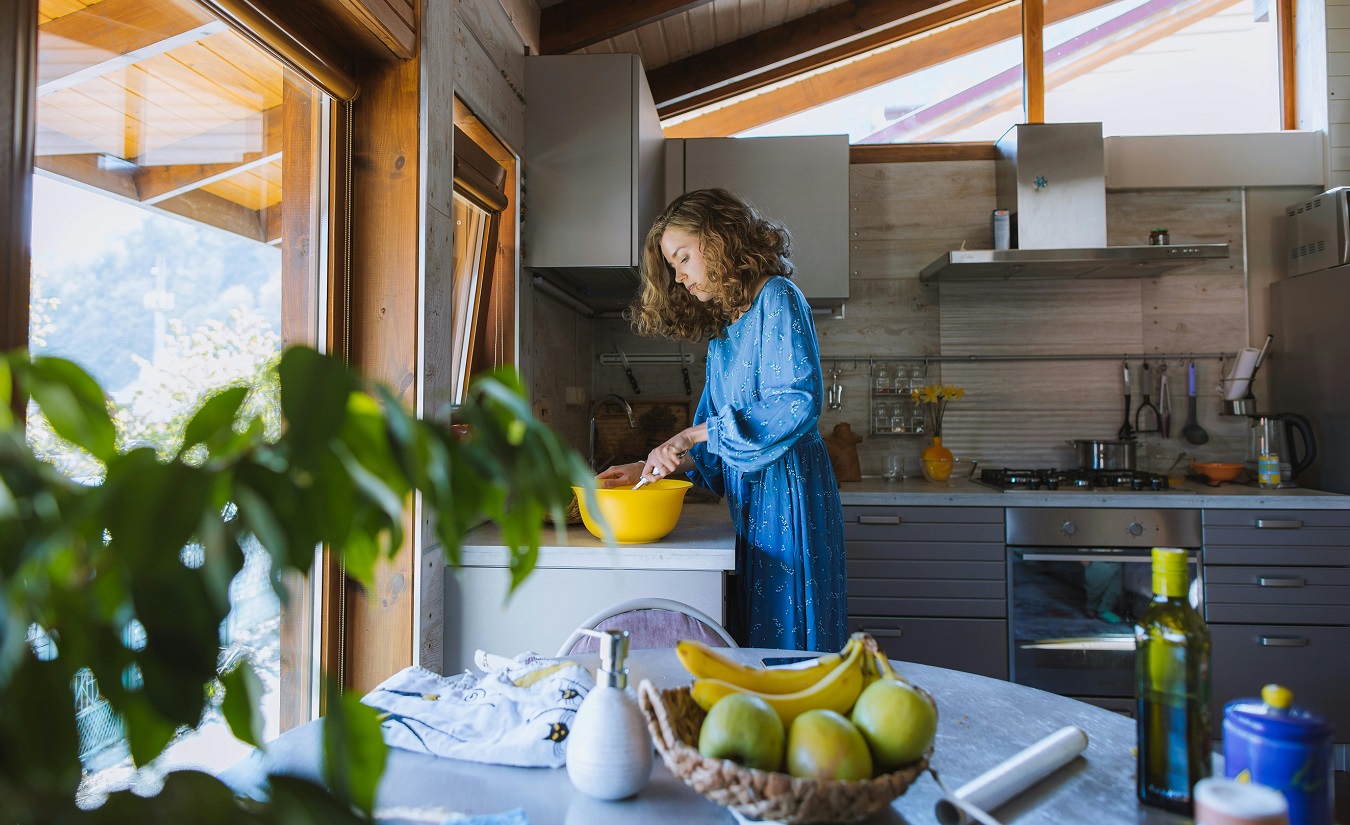 Eine Frau rührt in einer gelben Schüssel um, während sie in ihrer Küche steht. Sie trägt ein Nachthemd und hat eine schlanke Figur.