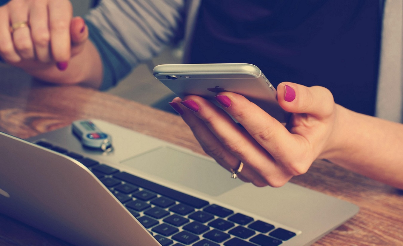 Eine Frau mit rot lackierten Fingernägeln hält ein Smartphone in der linken Hand und tippt mit der rechten Hand auf einem Laptop. Symbolbild für die MyTherapy Themenseite.