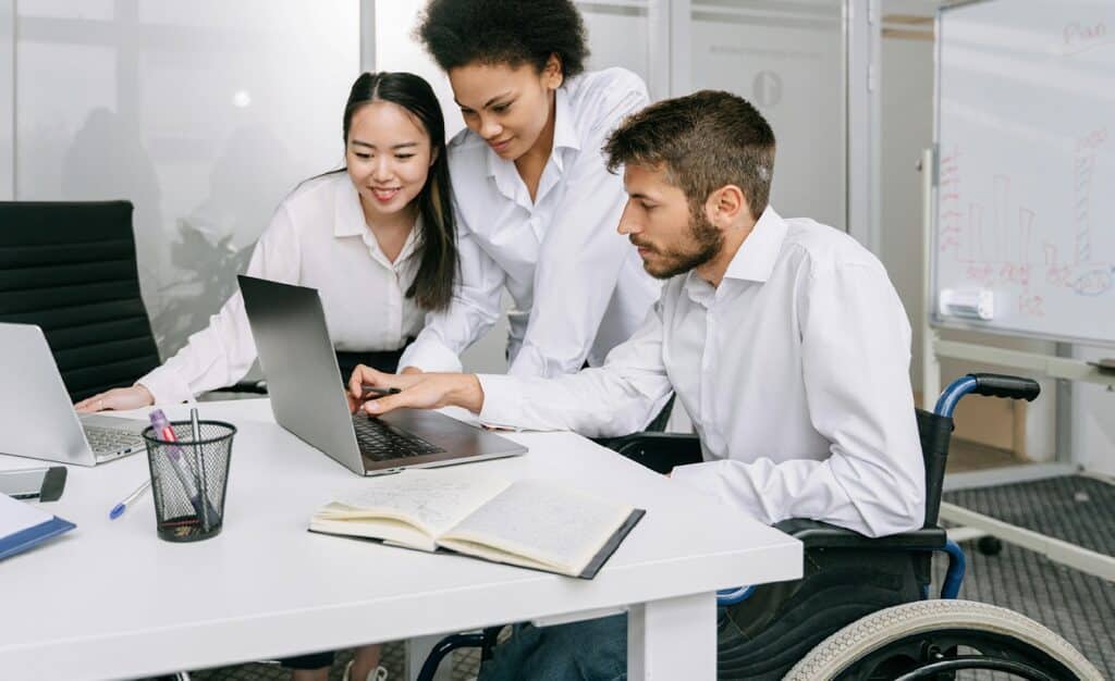 Drei Personen, darunter ein Mann im Rollstuhl, arbeiten gemeinsam an einem Laptop in einem Besprechungsraum. Im Hintergrund steht ein Flipchart. Alle tragen weiße Hemden und schwarze Hosen.