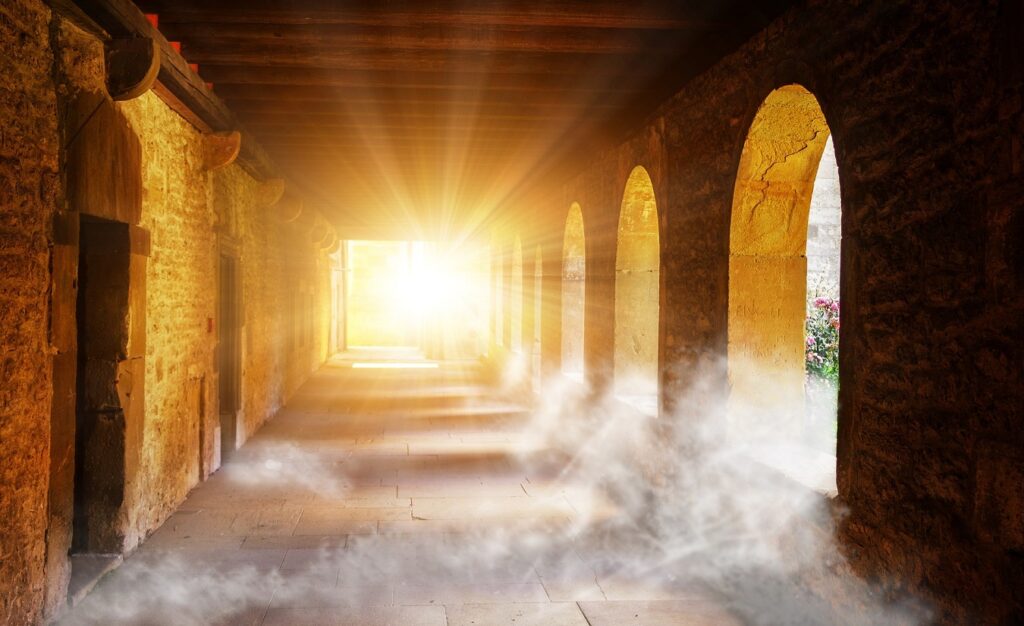 Ein nebelverhangener Gang in alten Klostermauern mit offenen Torbögen auf der rechten Seite. Am Ende des Gangs dringt Licht ein und verleiht dem Bild eine hoffnungsvolle Atmosphäre.