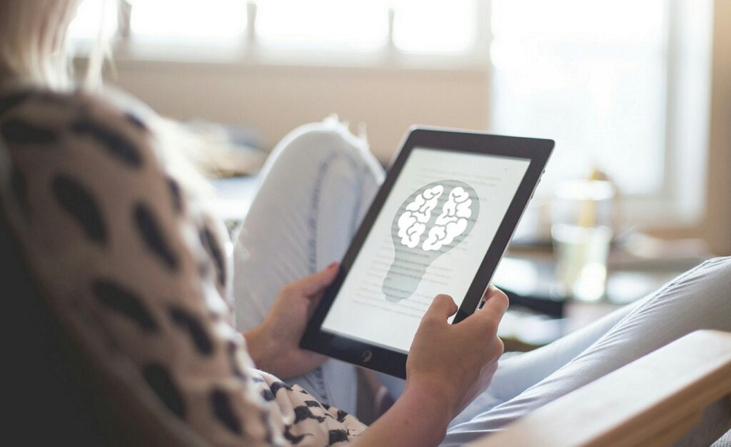 Frau sitzt im Wohnzimmer und hält ein Tablet mit einer Glühbirne und einer Gehirn-Illustration – Symbolbild für Informationen und Tipps für Menschen mit neu diagnostizierter Multipler Sklerose