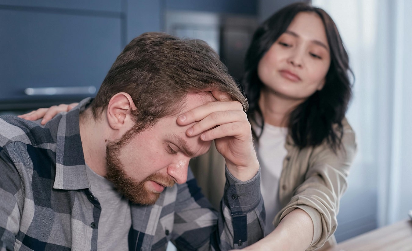 Ein Mann mittleren Alters hält sich schmerzgeplagt den Kopf, während eine Frau neben ihm steht und ihm tröstend beisteht – Symbolbild für Trigeminusneuralgie bei Multipler Sklerose.
